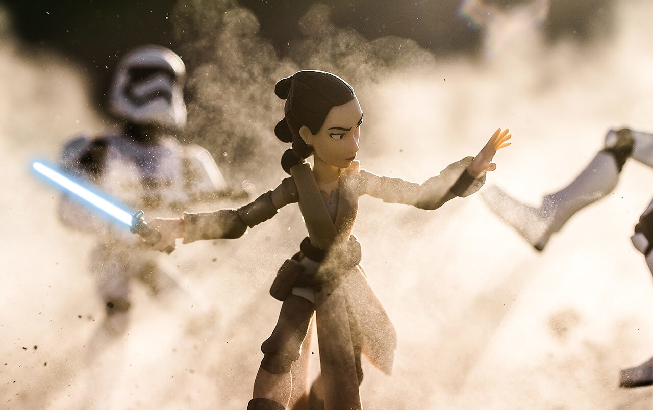 A Rey action figure is posed wielding a lighstaber while dirt and dust swirl around her and a stormtrooper action figure stands in the background.