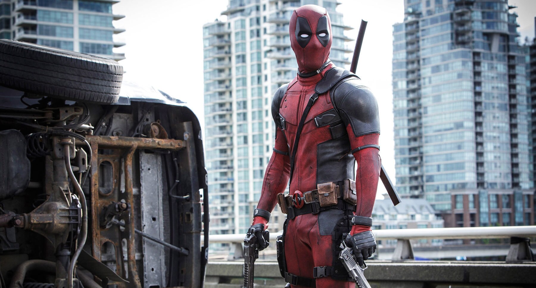 Ryan Reynolds (as Deadpool) with guns in his hands standing next to an overturned vehicle in "Deadpool"