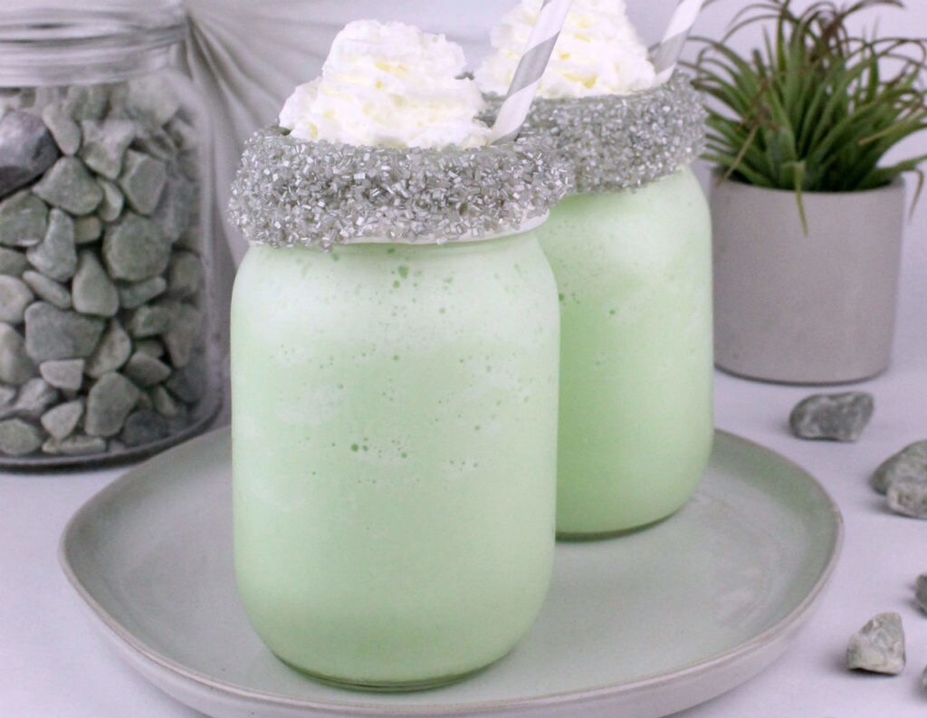Two green DIY thala-siren milkshakes, with silver sugar rims, whipped cream, and straws, in glass jars.