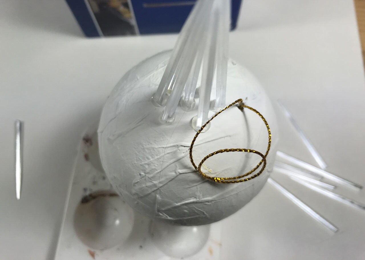 An unfinished, homemade vulptex Christmas ornament. Hot-glued clear toothpicks protrude from a bulb ornament painted white.
