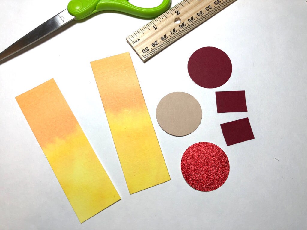 Two rectangular pieces of paper are painted yellow and orange to create the handmaiden's body while pieces of tan and red paper are cut into circles and squares to form her head and neck. This is a bookmark in progress.
