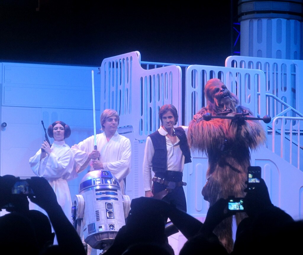 Fans take pictures of actors on stage dressed as Princess Leia, Luke Skywalker, Han Solo, and Chewbacca.