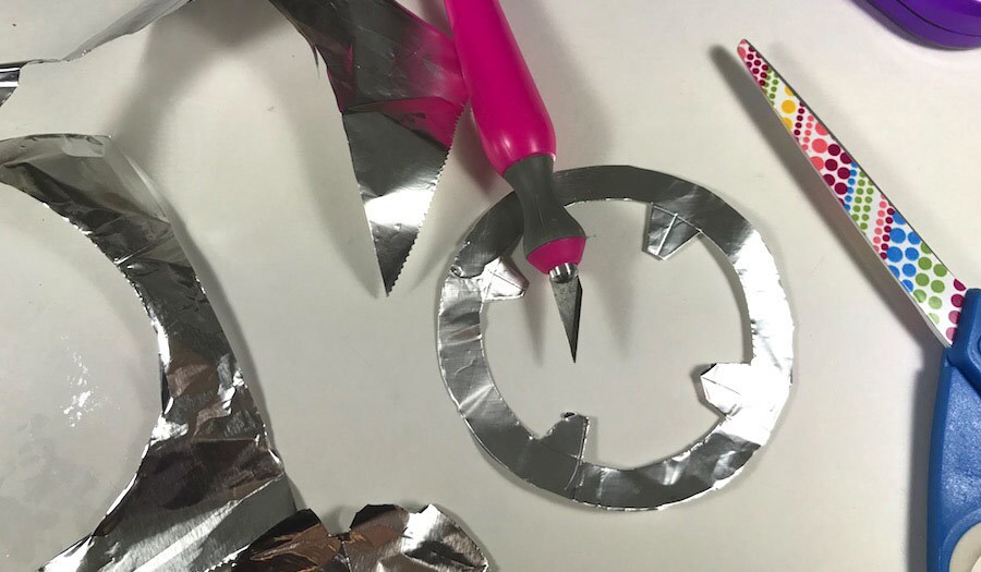 A craft knife and scissors lay atop a foil circle cut out.
