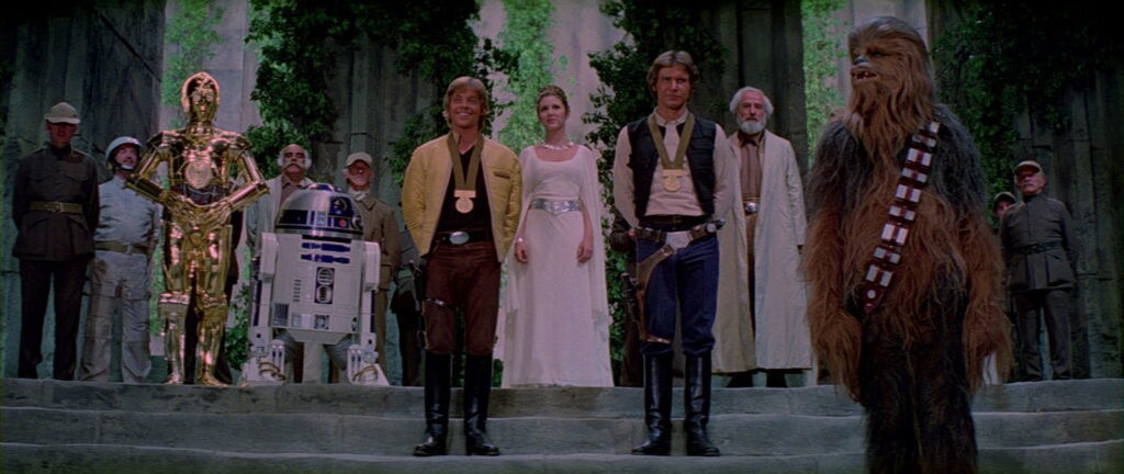 Luke and Han each wear medals while standing with C-3PO, R2-D2, Princess Leia, and Chewbacca during a victory ceremony in A New Hope.