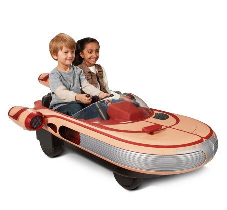 Two children sit in a small, drivable Landspeeder.