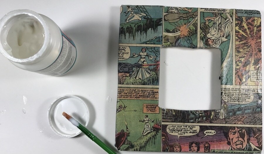 A bottle of glue, a paint brush, and a picture frame wrapped in comic book pages.