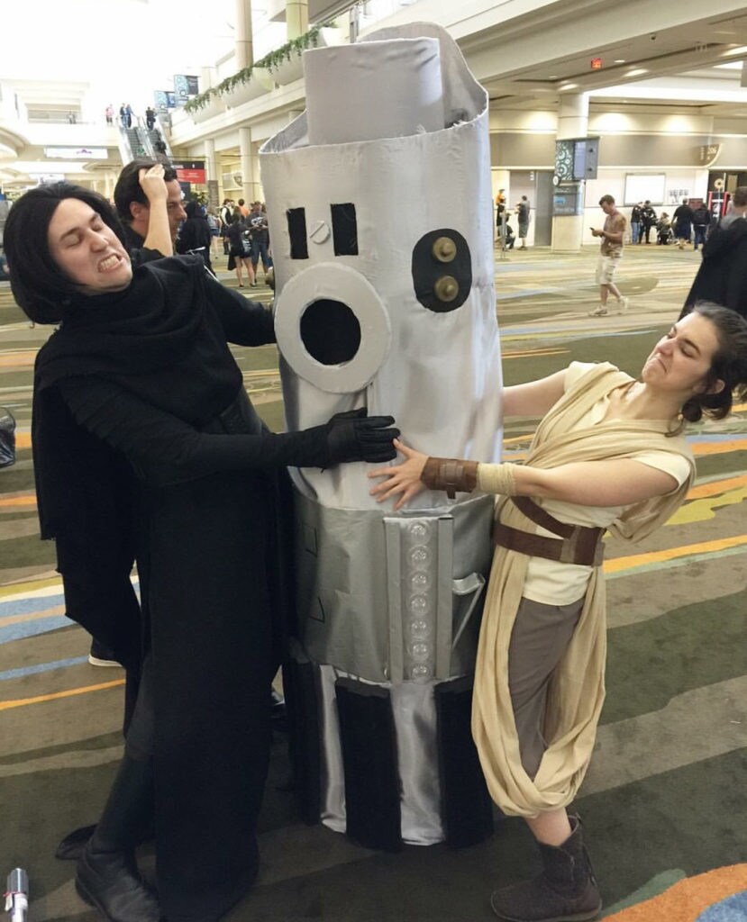 Rey and Kylo Ren cosplayers stand on either side of a third cosplayer dressed as Luke's lightsaber and pretend to fight to gain control of it.