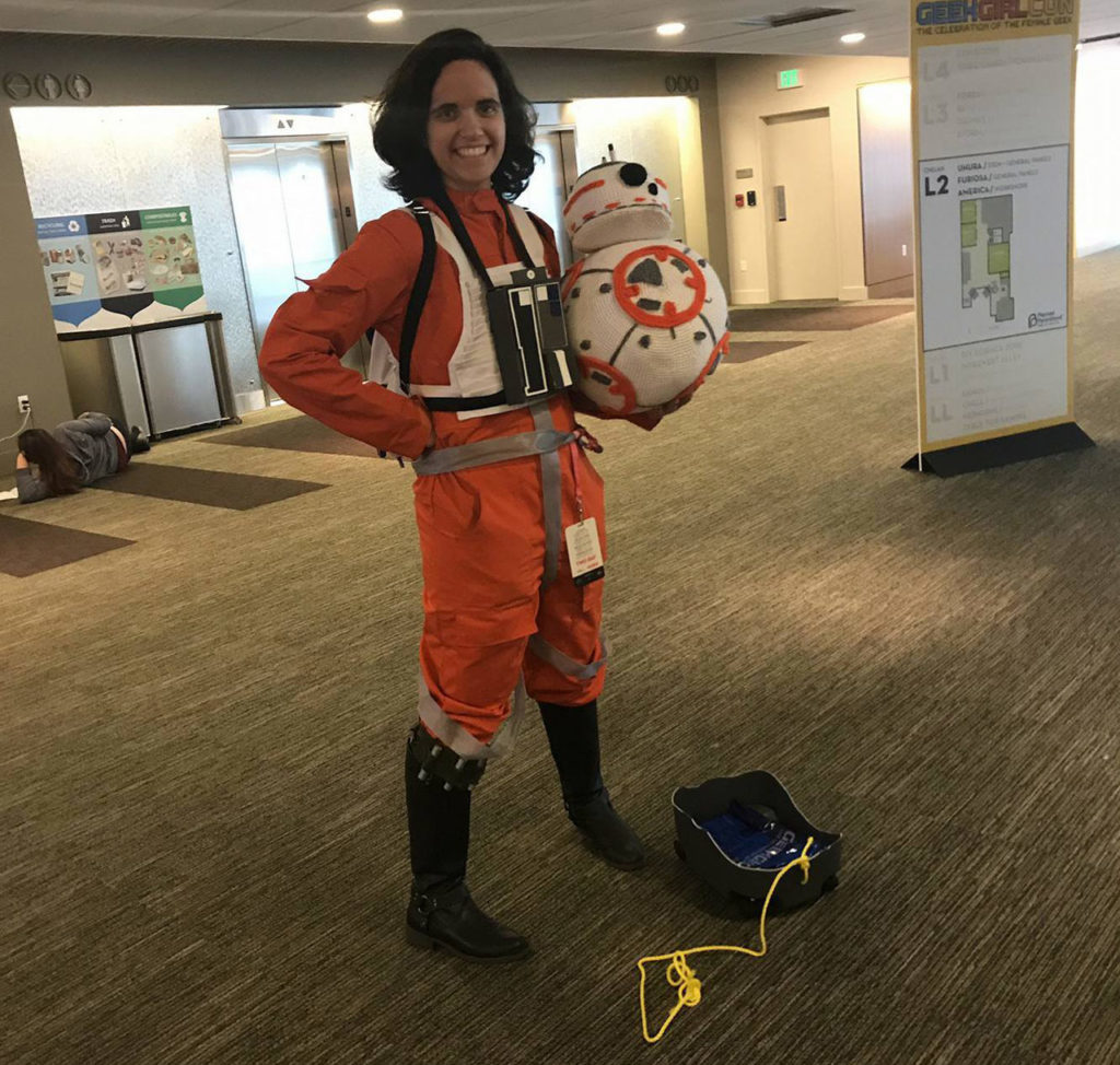 A cosplayer in Rebel pilot gear holds the crocheted BB-8 she created.