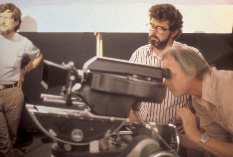 Visual effects artist, Dennis Muren, looks through a camera while George Lucas and another crew member stand nearby looking on.