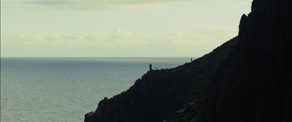 Seen from far away, Luke Skywalker watches Rey practice with her lightsaber on an island hillside in The Last Jedi.