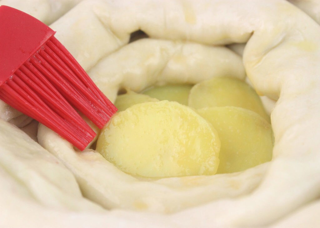 A pastry brush applies butter to dough wrapped around slices of potato.