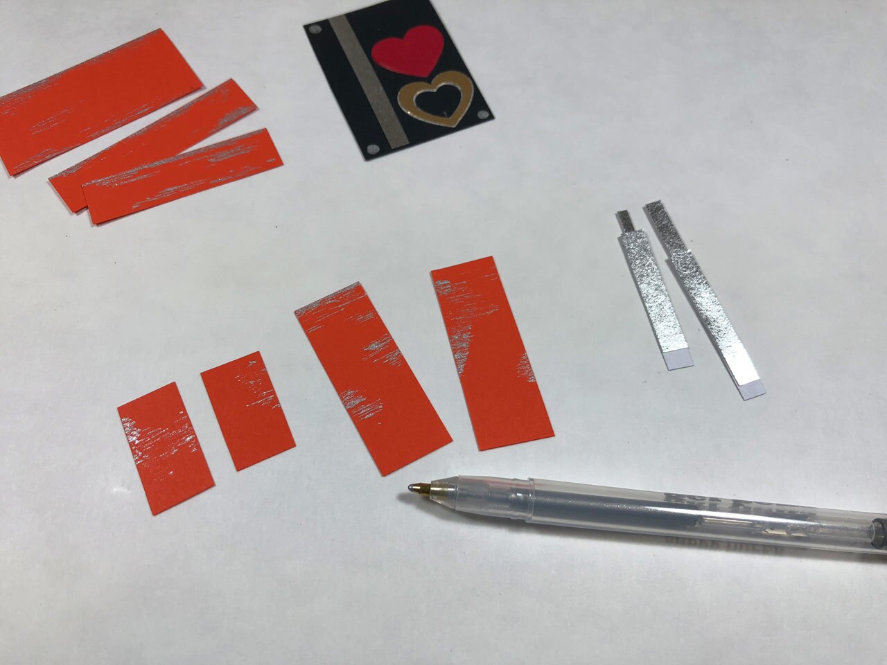 Strips of orange and silver metallic cardstock, black cardstock with red and gold paper hearts glued to it, and a silver gel pen. This is a Rose Tico-inspired Electro-Shock Prod Valentine's Day Card in progress.