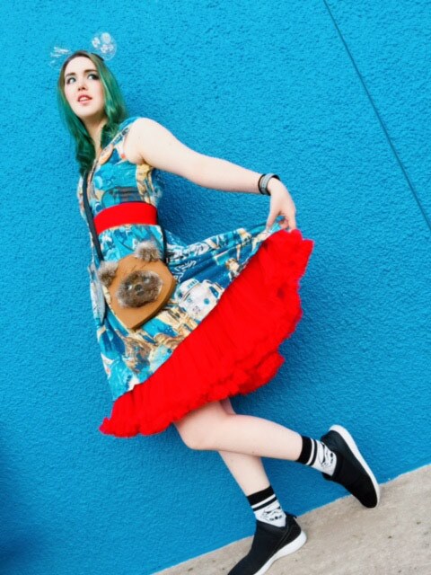 Cosplayer Moya Kojima shows off her Star Wars dress, Wookie bag, and stormtrooper socks in front of a blue wall.