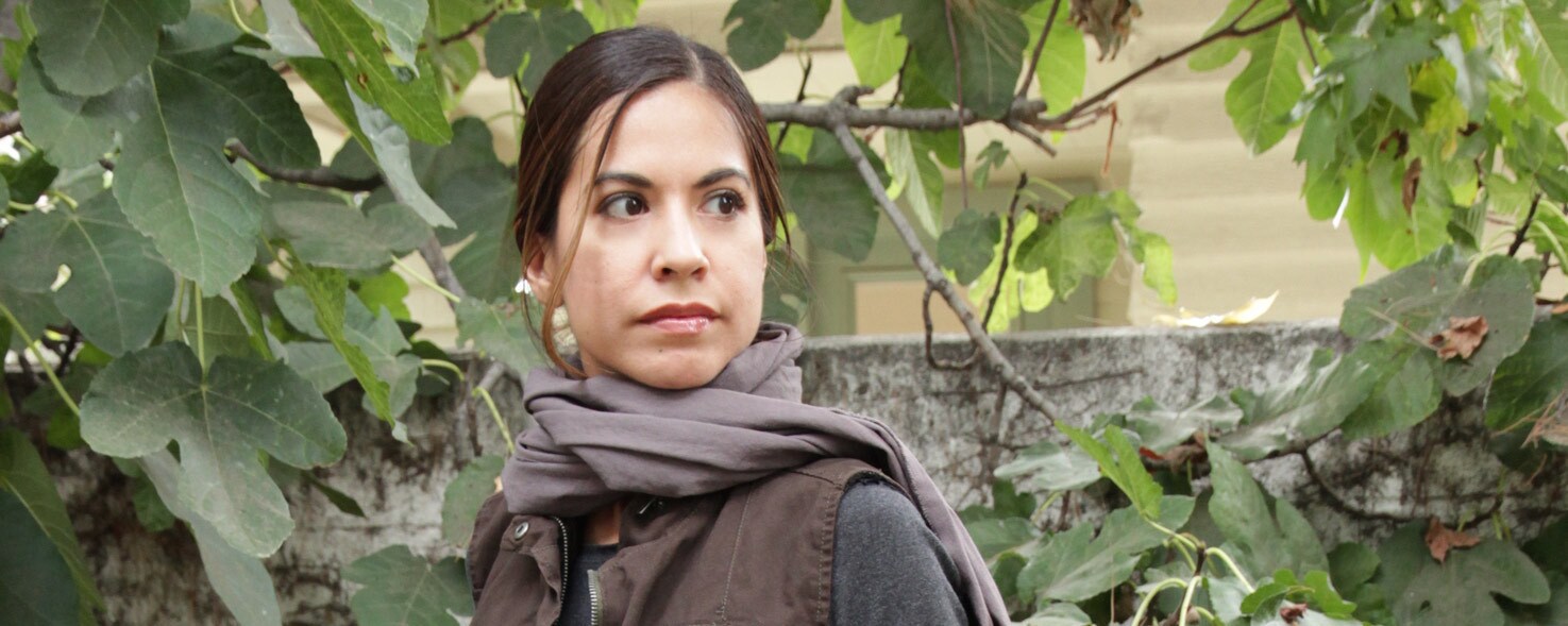 A female Star Wars fan models her do-it-yourself Jyn Erso Rogue One outfit which combines a long sleeve gray top, black flat-front pants, brown vest, and a gray scarf.