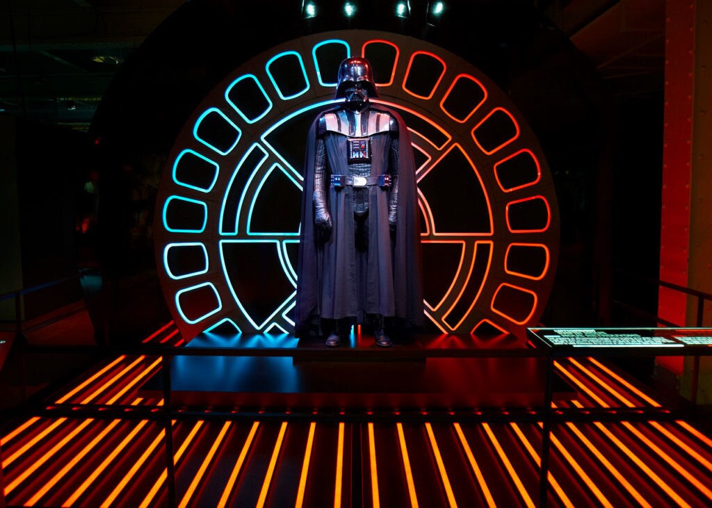 A Darth Vader costume against a background with blue and red lights on display at the Star Wars Identities exhibit.