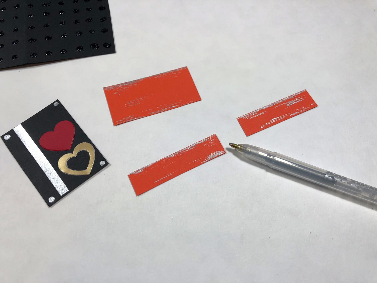 Strips of orange cardstock, black cardstock with red and gold hearts and a strip of silver metalllic cardstock glued to it, and a silver gel pen. This is a Rose Tico-inspired Electro-Shock Prod Valentine's Day Card in progress.