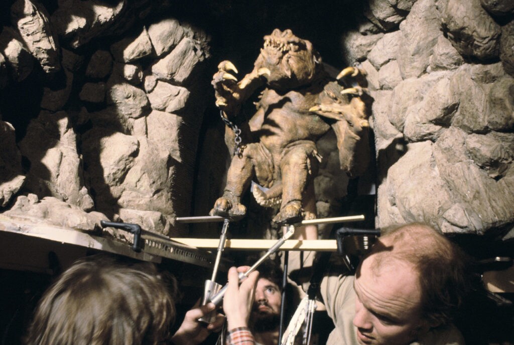 A three-person special effects team operates the reptilian rancor suit using various rods to move its body parts during the filming of Star Wars: Return of the Jedi.
