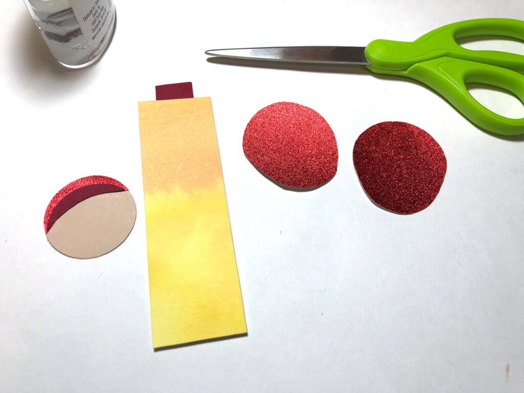 Round pieces of red crepe paper, a rectangular piece of yellow and orange water color paper, and a pair of green-handled scissors. This is a bookmark in progress.