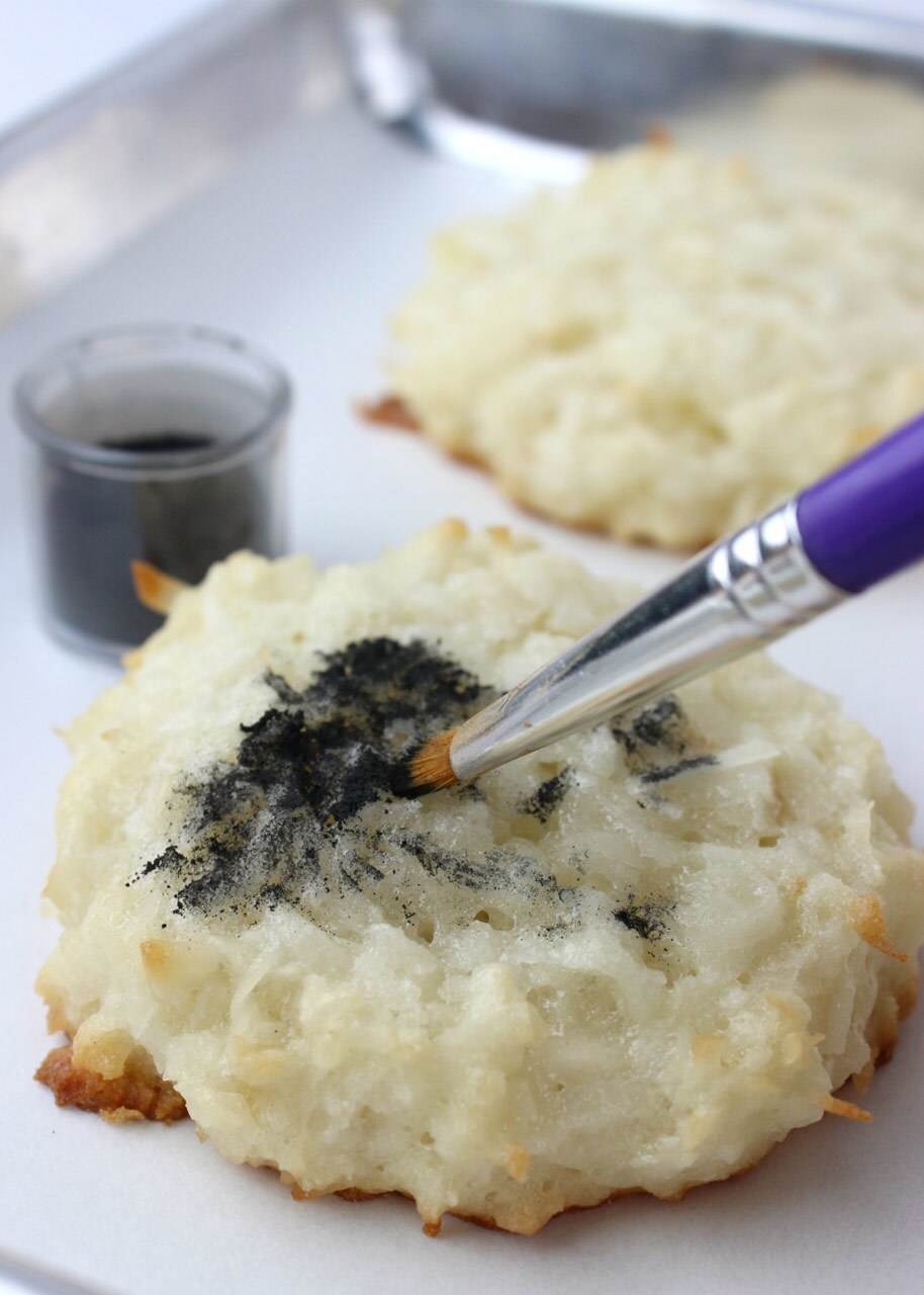 A kitchen brush paints black luster dust onto the center of an unfinished Muftak Macaroon.