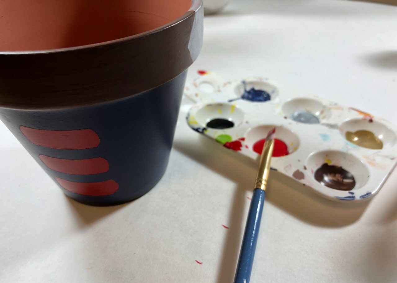 A terracotta pot painted blue with red stripes to make it Star Wars themed, with a tray of paint and a paintbrush next to it.