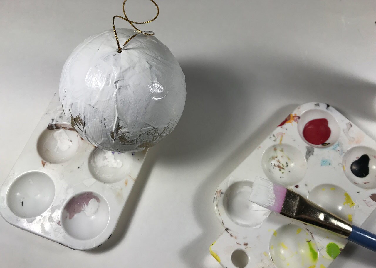 An unfinished, homemade vulptex Christmas ornament. A bulb ornament, painted white, dries atop a paint palette next to a second paint palette with a paintbrush.