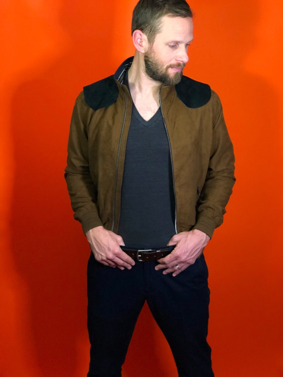 A male model wears his homemade Han Solo outfit featuring a brown suede bomber jacket, charcoal gray v-neck tee, navy pants, and a brown belt.