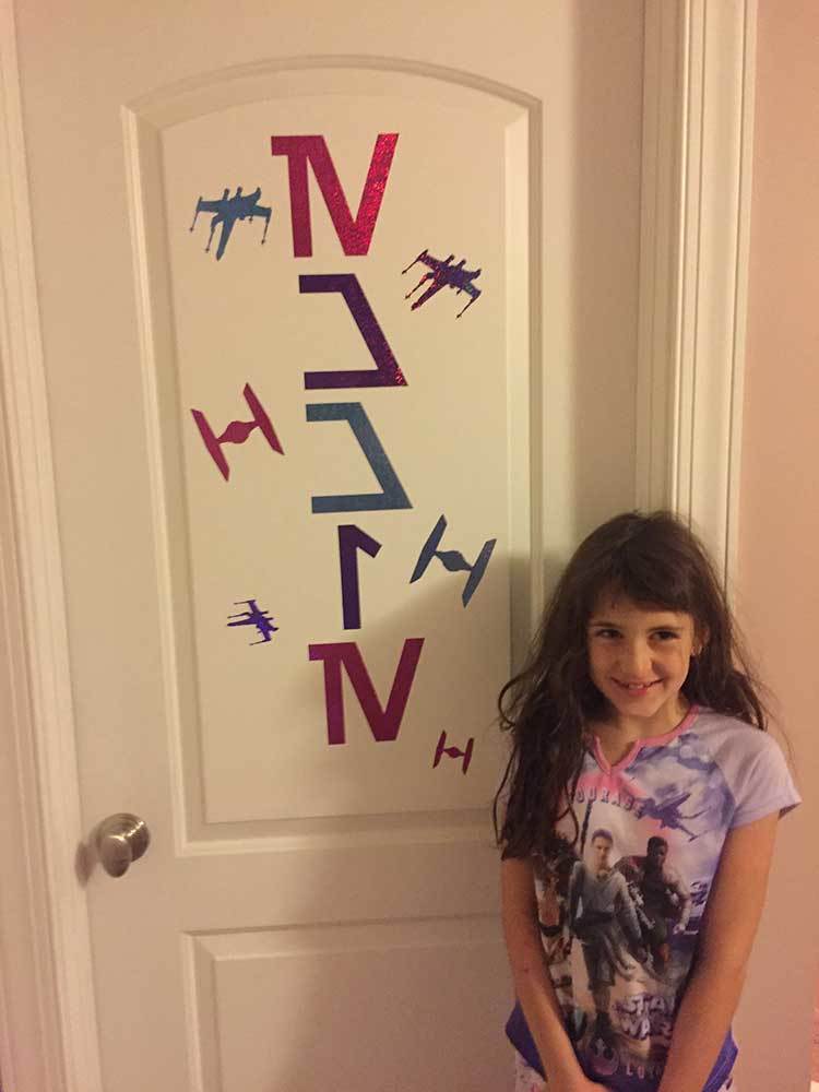 A Star Wars super-fan stands next to a door. Her name, Emmie, is spelled out in pink and purple decals in the classic Star Wars Language: Aurebesh.