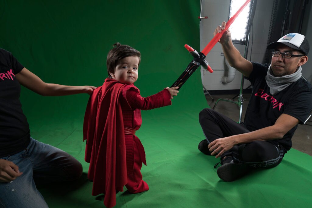 Josh Rossi and his son, who is holding a red lightsaber, pose in front of a green screen.