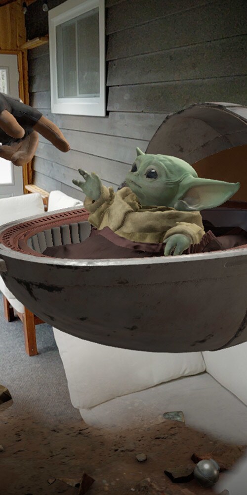 A finger reaches out, pointing at Grogu in a floating basinet, in an augmented reality experience.