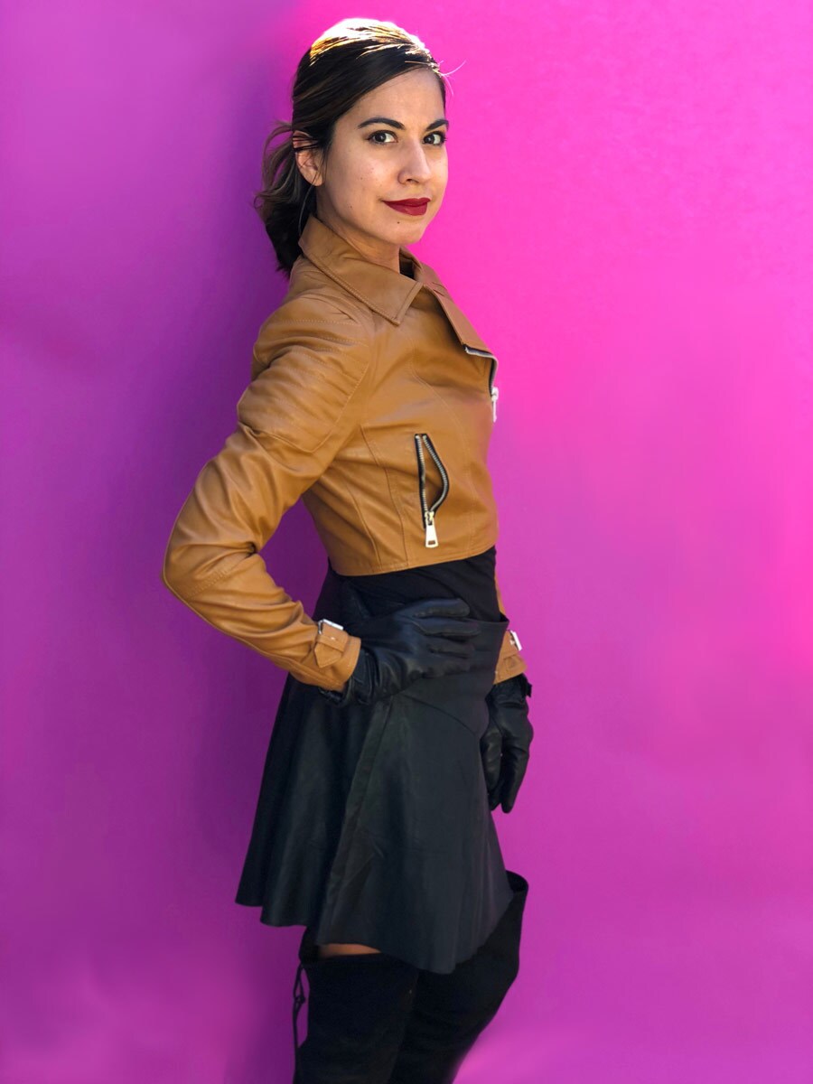 A female model wears her homemade Qi'ra outfit featuring a tan moto jacket, black leather skirt, black boots, and black gloves.