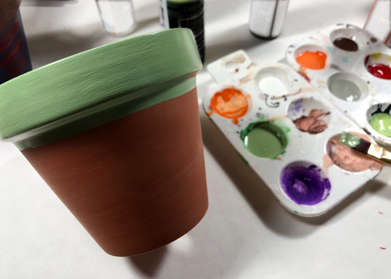 A terracotta pot with the top painted green next to a paintbrush and a tray of paint.