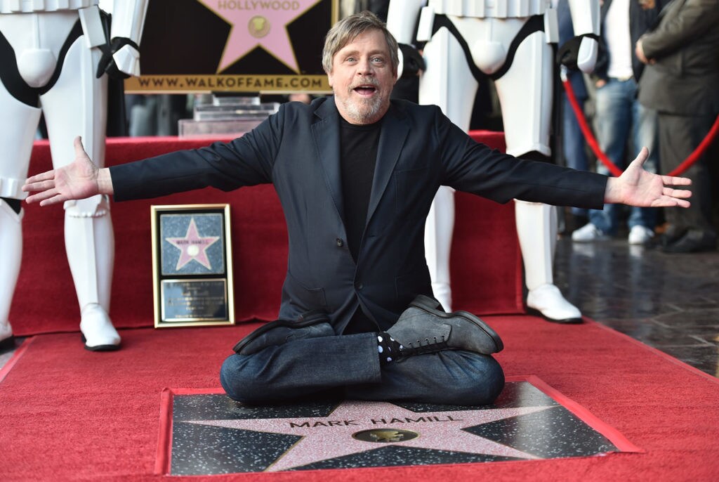 Mark Hamill sits on a red carpet with his arms wide open as he displays his star on the Hollywood Walk of Fame.