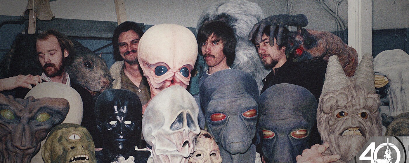 The heads of the various aliens seen in the Mos Eisley Cantina in A New Hope sit on a workshop table and the artists who created them stand behind.