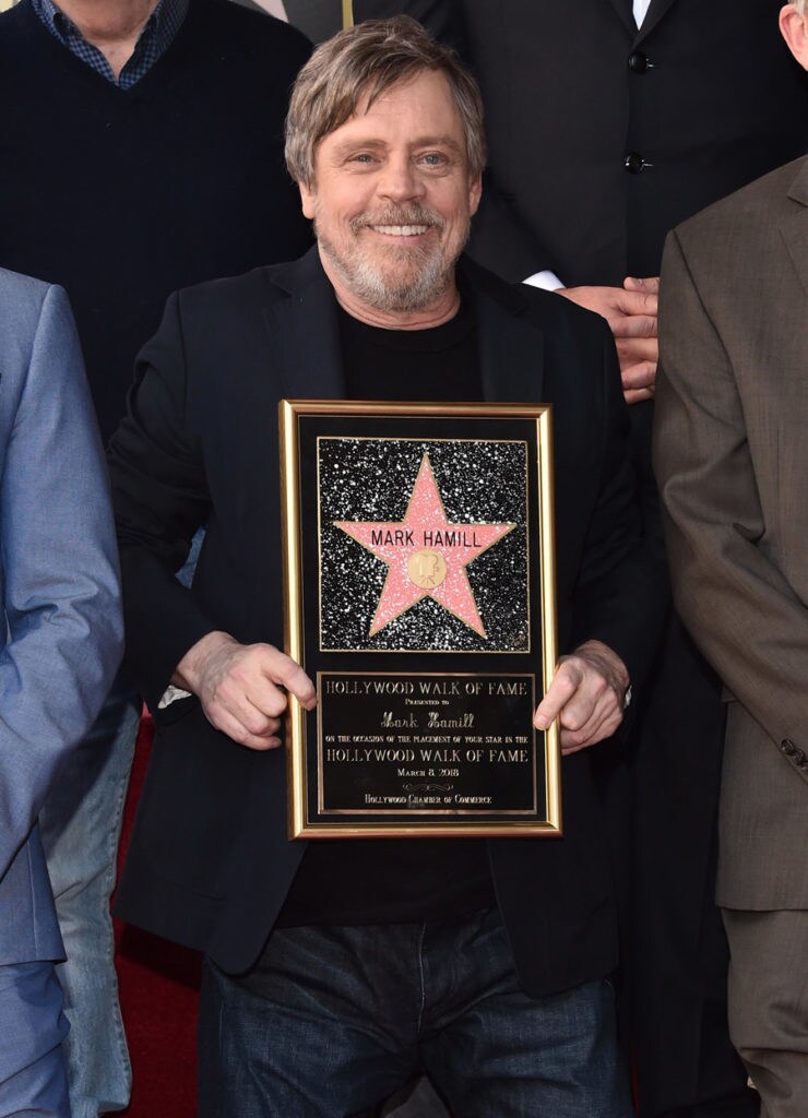 Mark Hamill holds a plaque showing a replica of his star on the Hollywood Walk of Fame. The bottom of the plaque shows text commemorating the occasion.