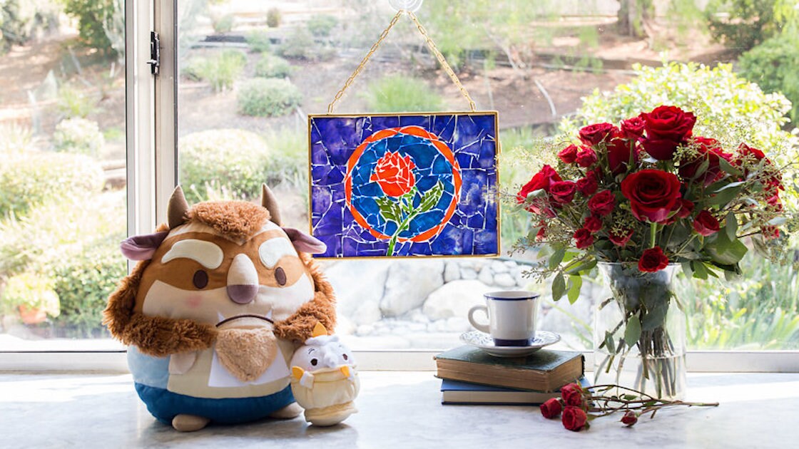 The stained glass rose from Beauty and the Beast crafted from pieces of dyed pasta hangs in a window behind a stuffed Ufufy Beast and Lumiere and an arrangement of red roses.