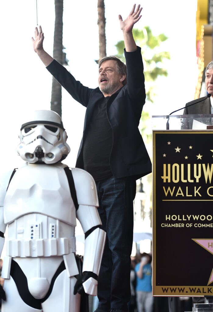 Mark Hamill raises his hands high in the air while a stormtrooper stands in front of him at a ceremony honoring him with a star on the Hollywood Walk of Fame.