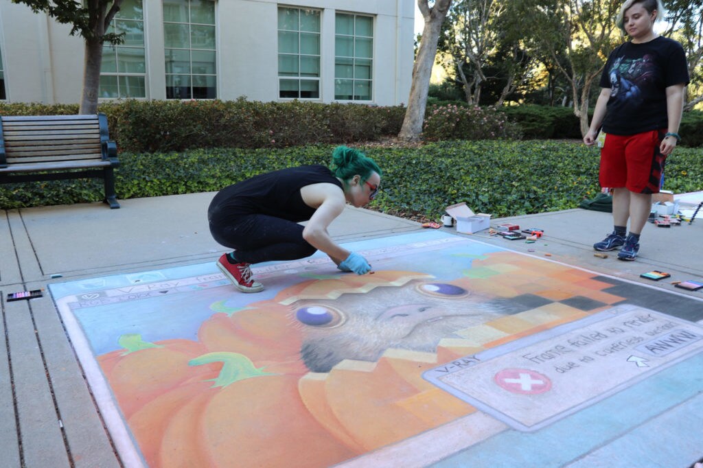 Two artists work on sidewalk chalk art of a computer screen displaying a porg poking its head out of a pumpkin in a pumpkin patch.