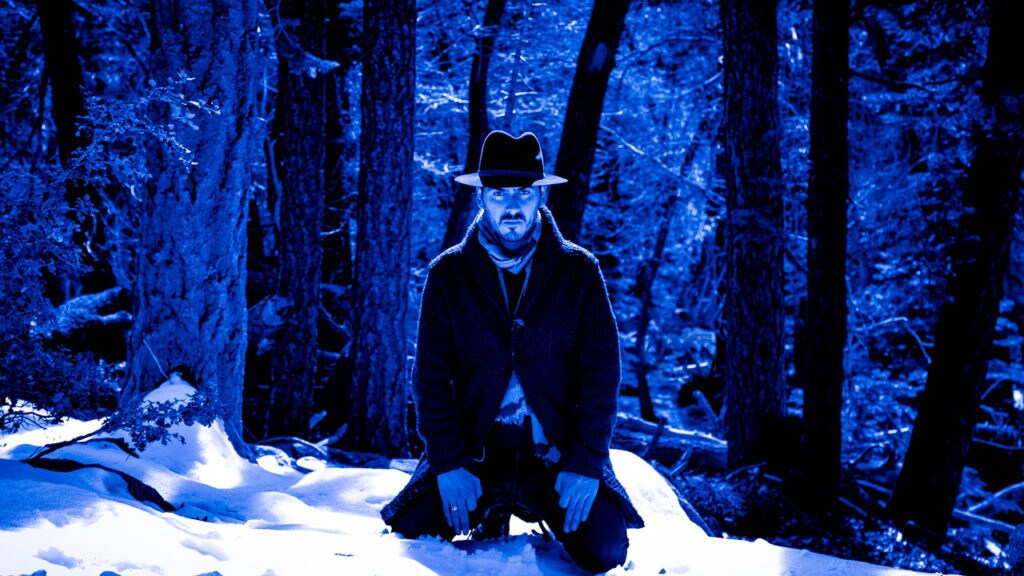 Dhani Harrison in a hat and coat in the forrest in the snow.
