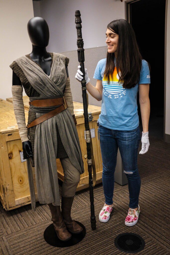Lucasfilm archivist Madlyn Burkett holds Rey's staff from The Force Awakens while standing beside a mannequin wearing one of Rey's outfits.