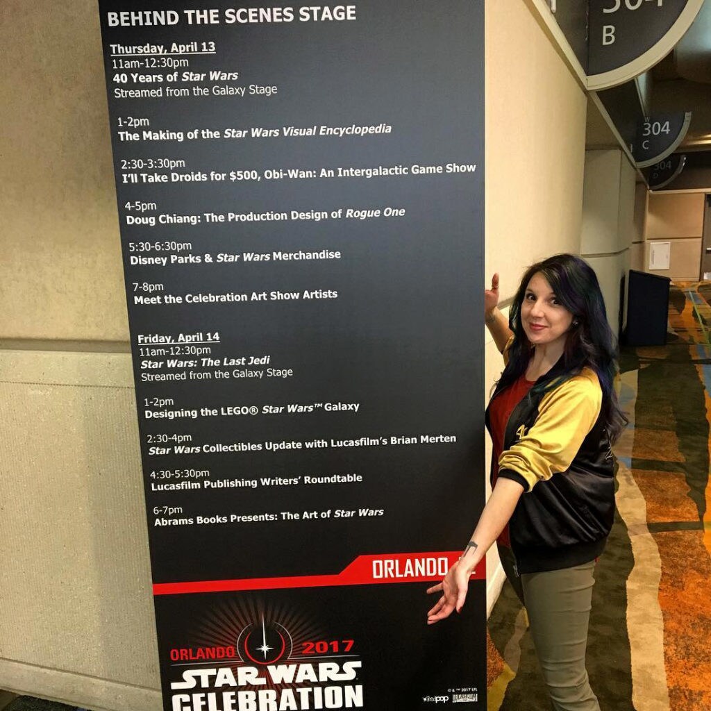 Host and author Amy Ratcliffe stands beside a large sign detailing the schedule of events at Star Wars Celebration Orlando.