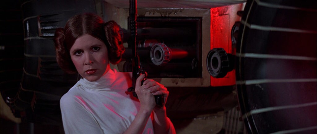 Princess Leia, in a simple white gown and hair in two buns, holds a blaster in Star Wars: A New Hope.