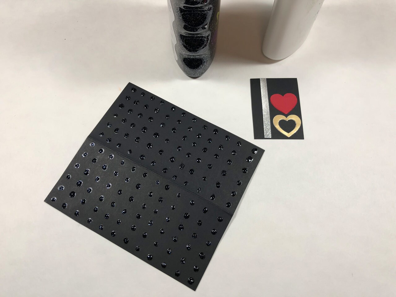 A square piece of black cardstock with rows of black glitter glue dots, and a smaller piece of black cardstock with a strip of silver metallic cardstock and red and gold paper hearts. This is a Rose Tico-inspired Electro-Shock Prod Valentine's Day Card in progress.