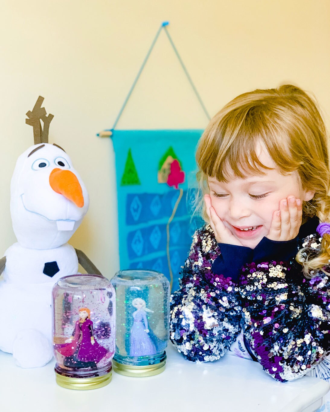 A little girl and her Olaf doll pose next to two homemade snow globes made from mason jars, featuring Anna and Elsa.