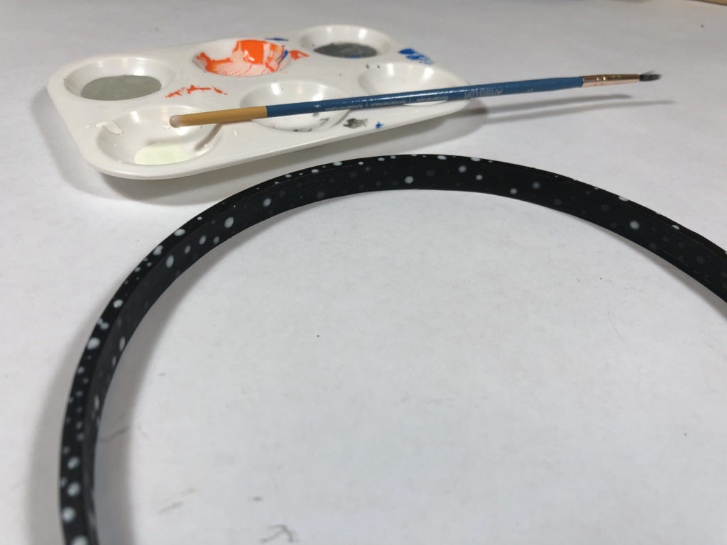 An embroidery hoop painted black with glow-in-the-dark specks next to a paint palette and paintbrush.