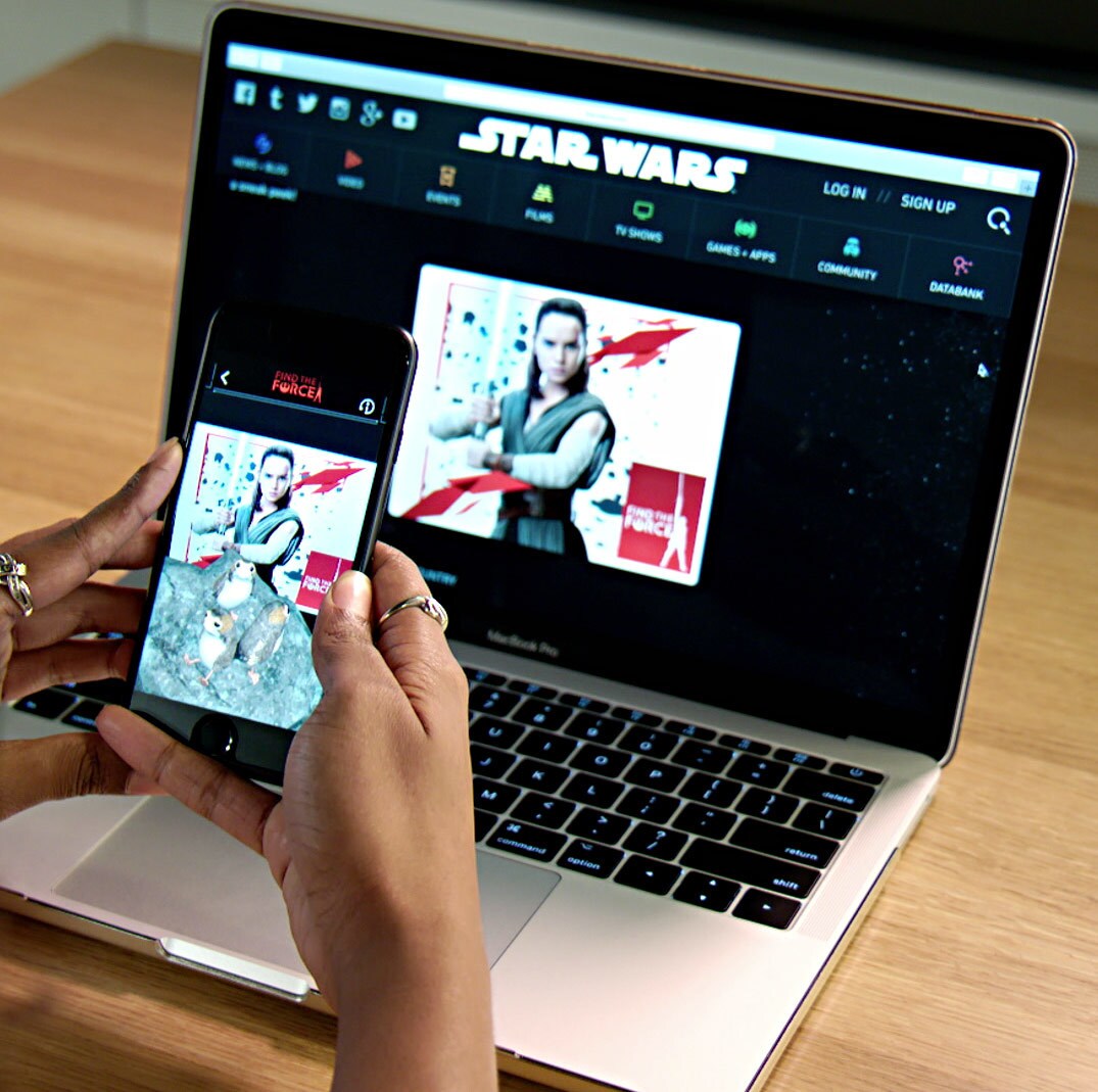 An augmented reality version of Rey with Porgs appears on a person's phone in front of a picture of Rey on a laptop.