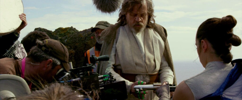 A behind-the-scenes photo from The Last Jedi shows Rey handing Luke his lightsaber while a film crew captures the moment.