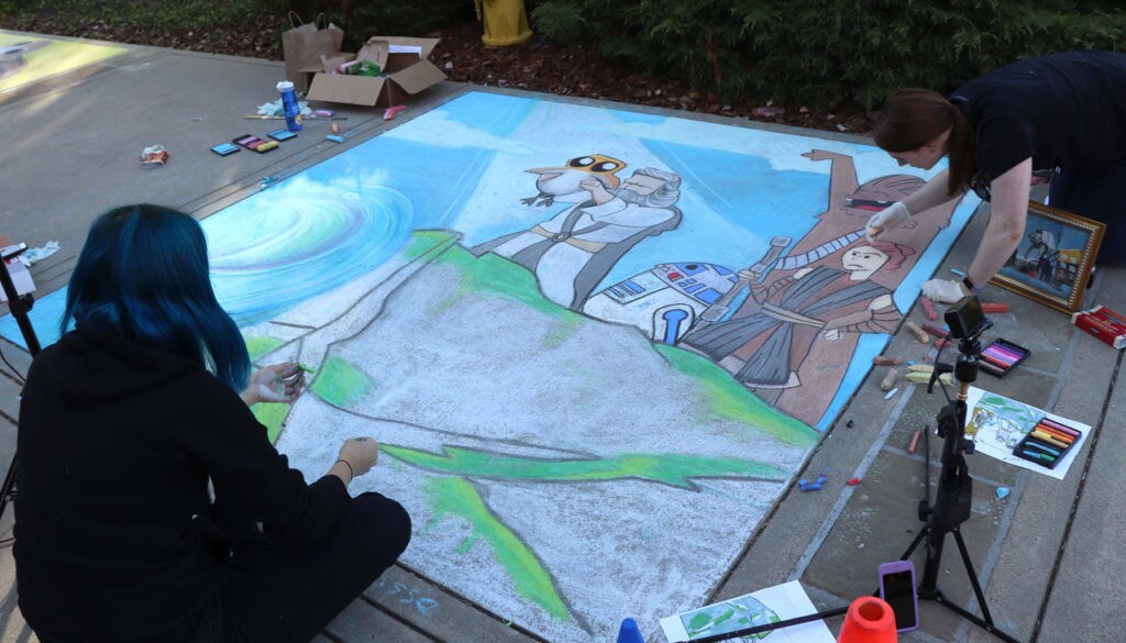 An artist works on sidewalk art inspired by the Lion King, with Rey, Chewbacca, and R2-D2 looking on as Luke Skywalker holds a Porg on a cliff.
