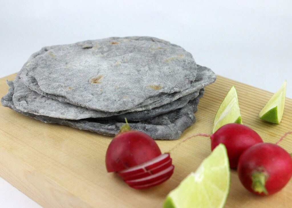 A stack of grey tortillas on a wooden board with lime wedges and radishes.