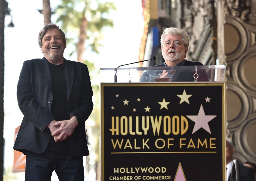 Mark Hamill laughs while standing beside George Lucas who speaks at a podium at a ceremony honoring Mark Hamill with a star on the Hollywood Walk of Fame.