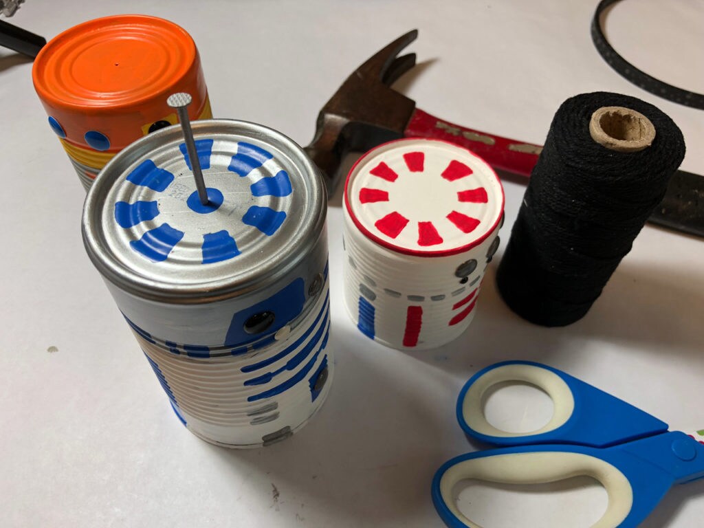 Tin cans painted to look like Chopper, R2-D2, and R5-D4 next to a hammer, black string, and a pair of scissors.